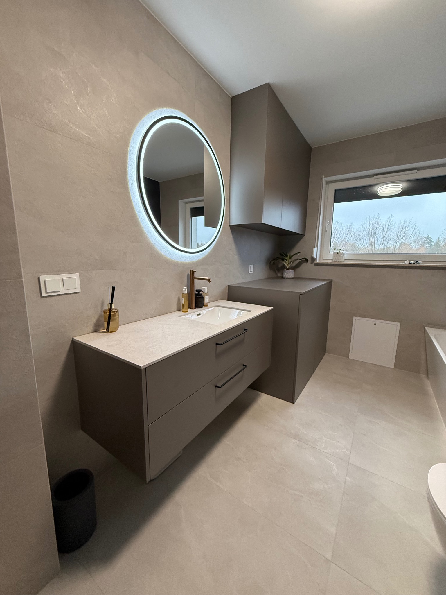 Nowoczesna łazienka z szarą szafką pod umywalką, okrągłym lustrem z podświetleniem LED i wiszącą szafką. Minimalistyczny design, neutralna kolorystyka.