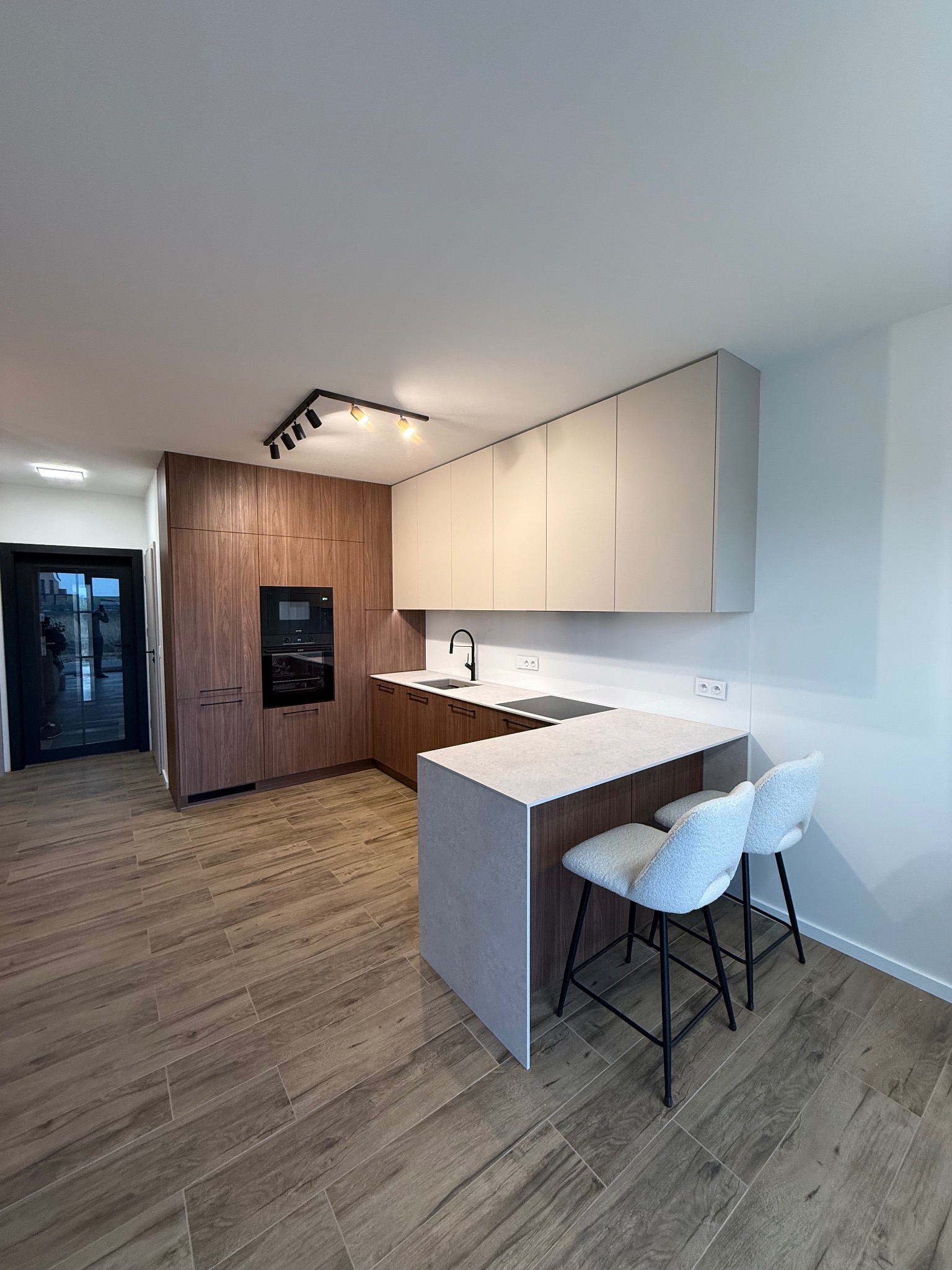 Nowoczesna kuchnia z drewnianymi frontami i wyspą z szarym blatem. Dwa białe krzesła barowe. Oświetlenie punktowe. Podłoga imitująca drewno. Minimalistyczny design.