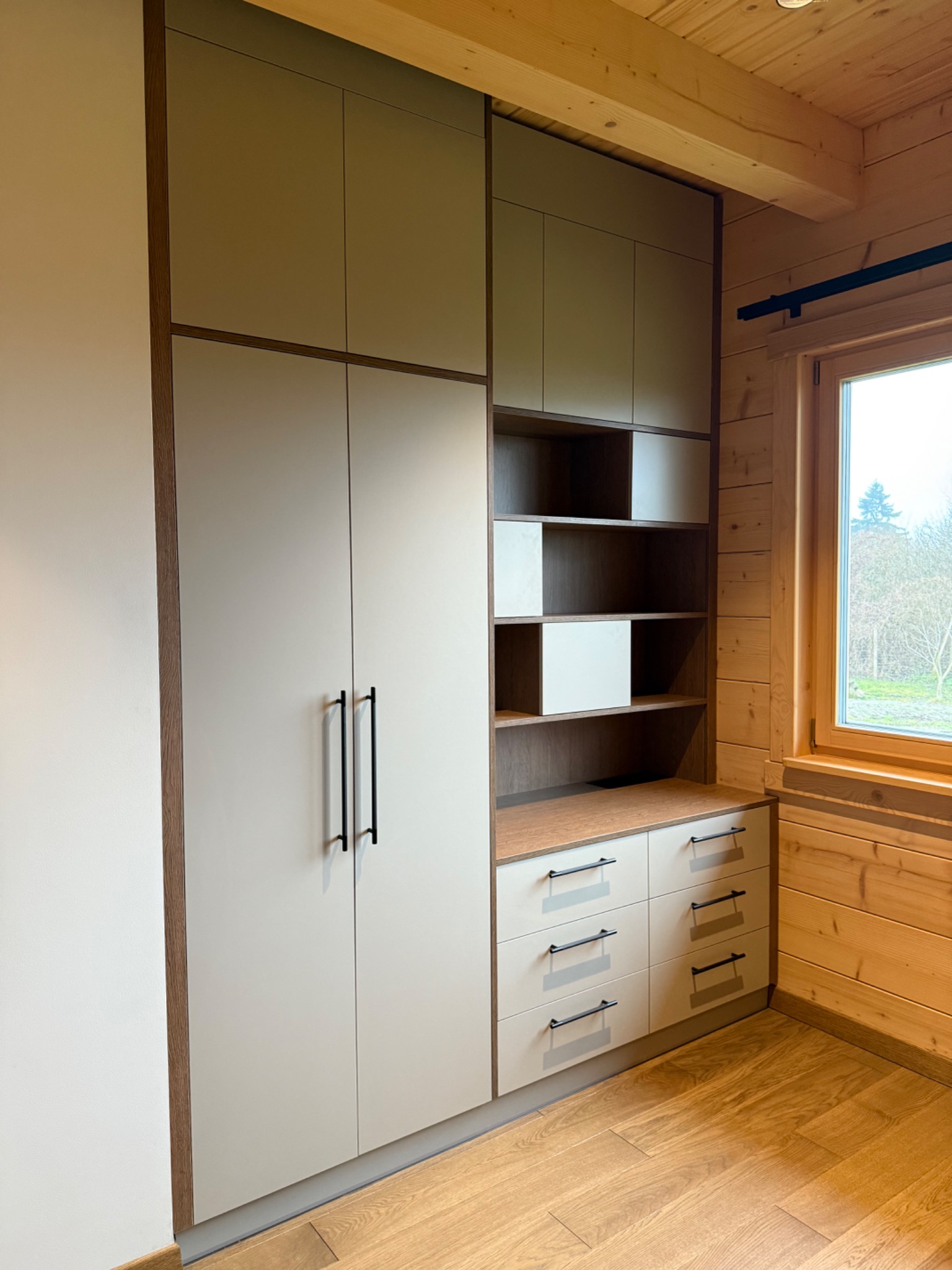 Jasna szafa wnękowa z szufladami i półkami, czarne uchwyty, drewniane wykończenie, wnętrze domu z drewnianymi ścianami i podłogą, widok na okno.