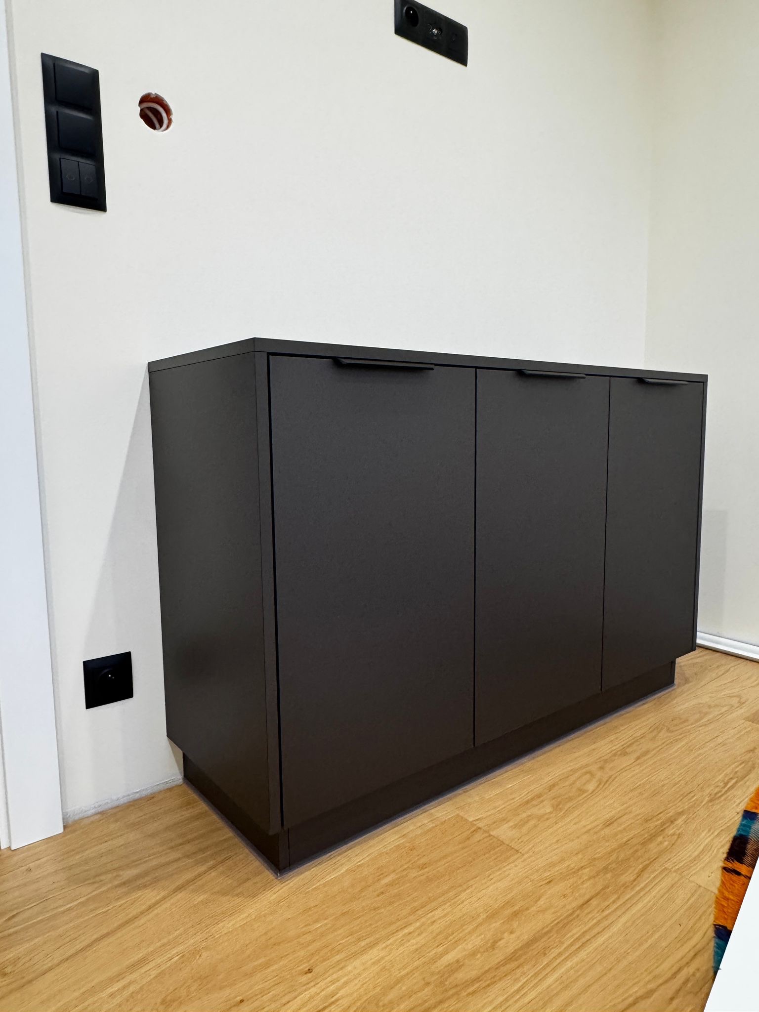 Ciemna, matowa szafka pod zabudowę z trzema drzwiczkami i minimalistycznymi uchwytami, na tle białej ściany z widocznymi gniazdkami i przełącznikami.