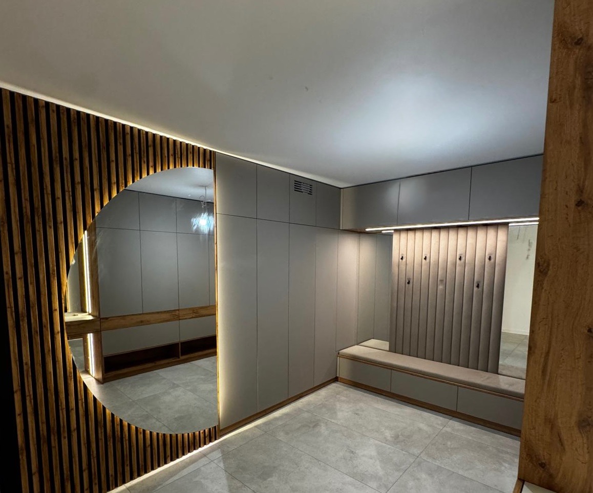 Nowoczesna szafa wnękowa z siedziskiem i lustrem w kształcie półkola, podświetlona listwami LED. Szara kolorystyka, drewniane akcenty i minimalistyczny design.