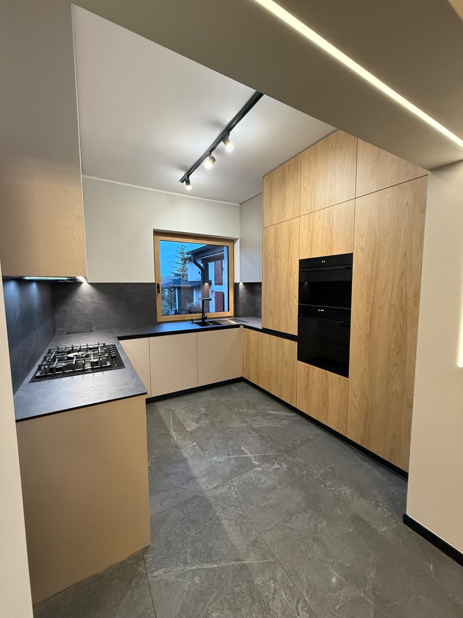 Nowoczesna kuchnia z drewnianymi frontami szafek, czarnym blatem i szarą podłogą. Oświetlenie punktowe i okno z widokiem na zewnątrz. Minimalistyczny design.