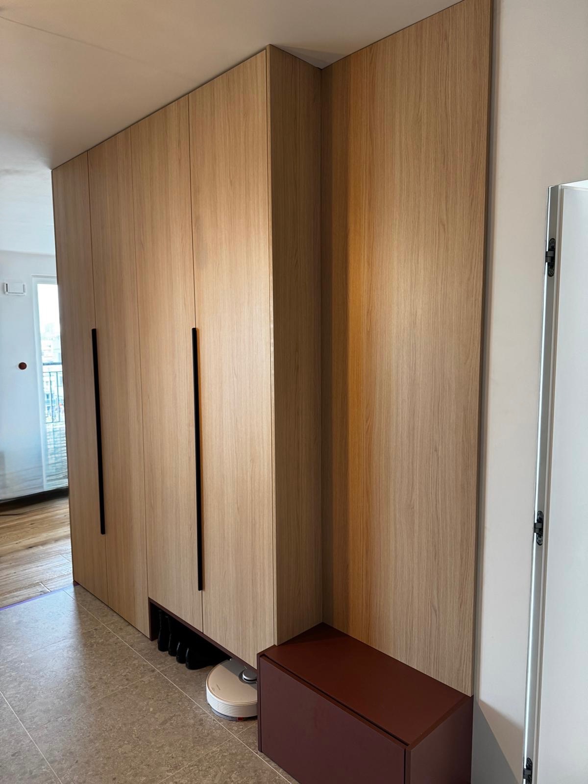 Jasna, drewniana szafa wnękowa z czarnymi uchwytami, dopasowana do ściany. Widoczny fragment podłogi i listwy przypodłogowej. Pod szafą robot sprzątający.