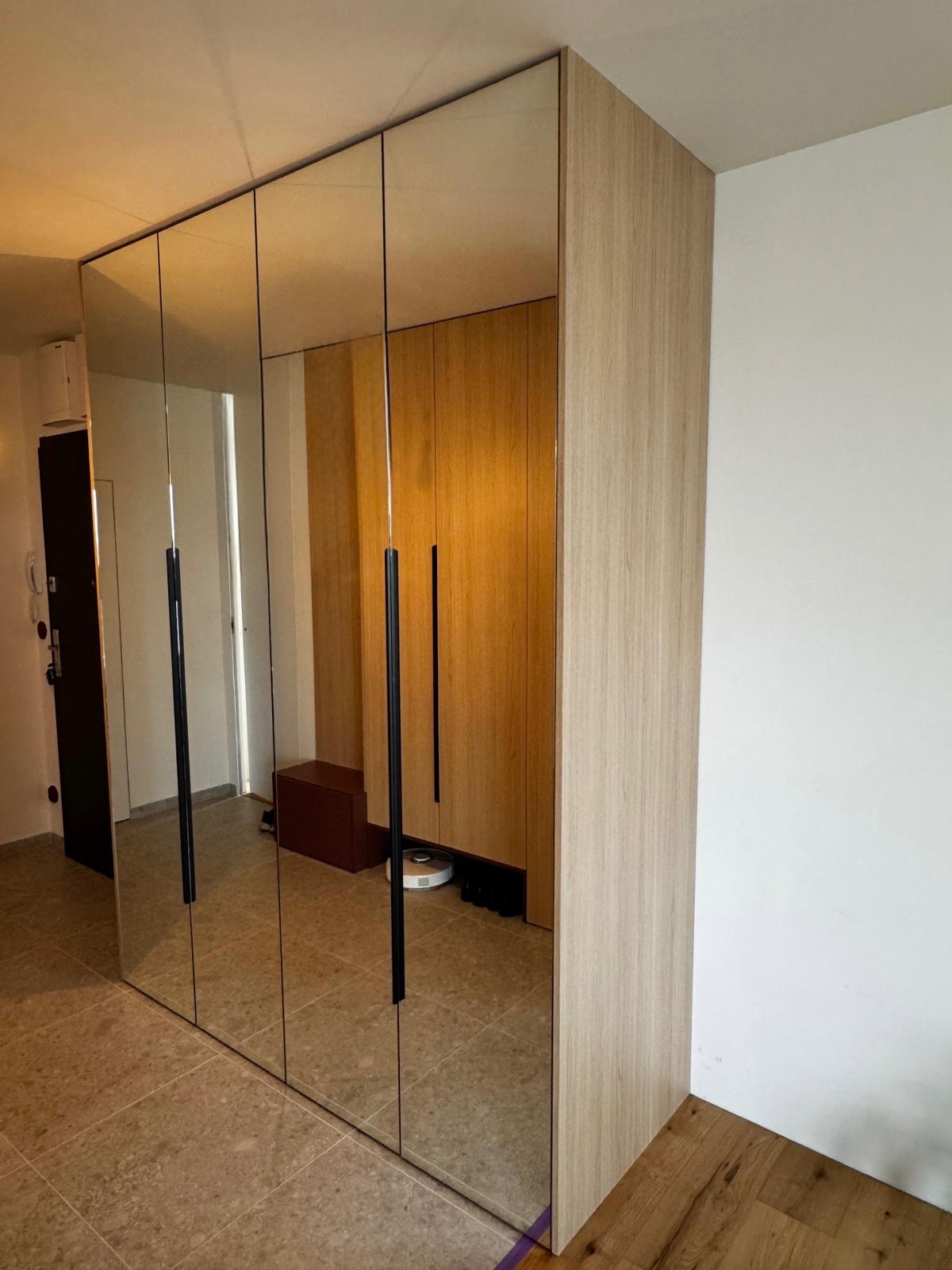 Szafa wnękowa z lustrzanymi drzwiami, czarne, długie uchwyty, jasne drewno na bocznej ściance, widoczne odbicie wnętrza mieszkania.