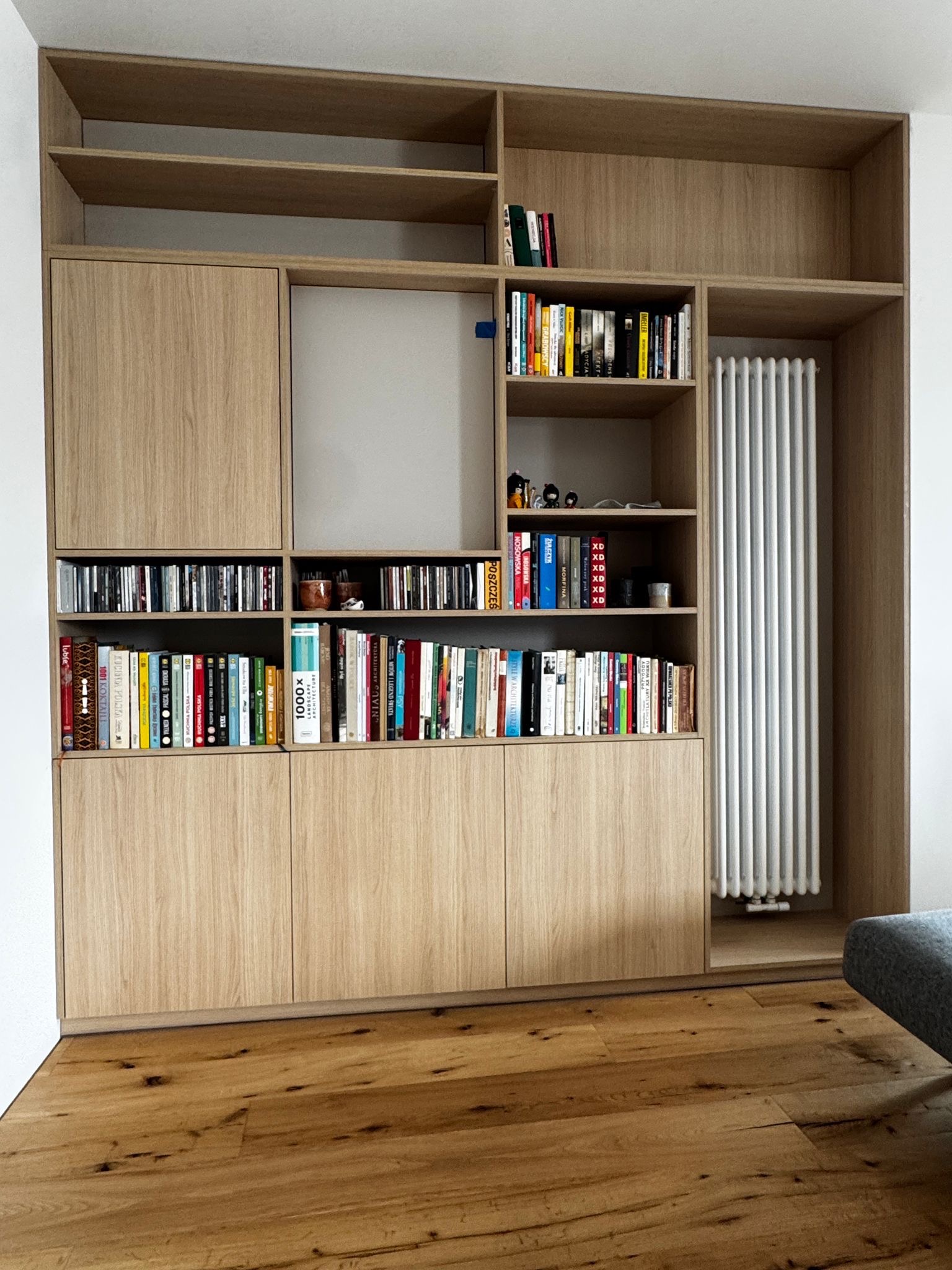 Drewniana zabudowa biblioteczna na wymiar z półkami i szafkami, wypełniona książkami i płytami CD. Grzejnik wkomponowany w konstrukcję. Parkiet na podłodze.