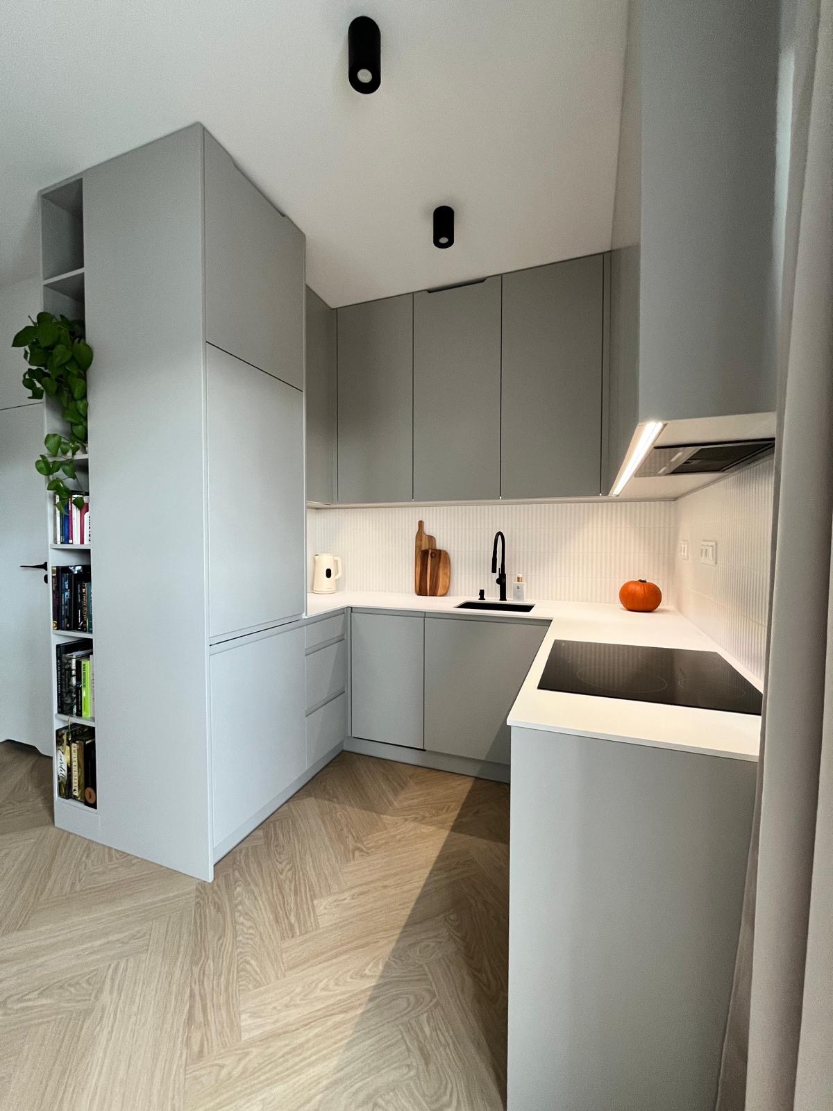 Jasnoszara kuchnia w nowoczesnym stylu z drewnianą podłogą w jodełkę. Na blacie czarna bateria, deski do krojenia i dynia. Dodatkowa półka na książki i rośliny.