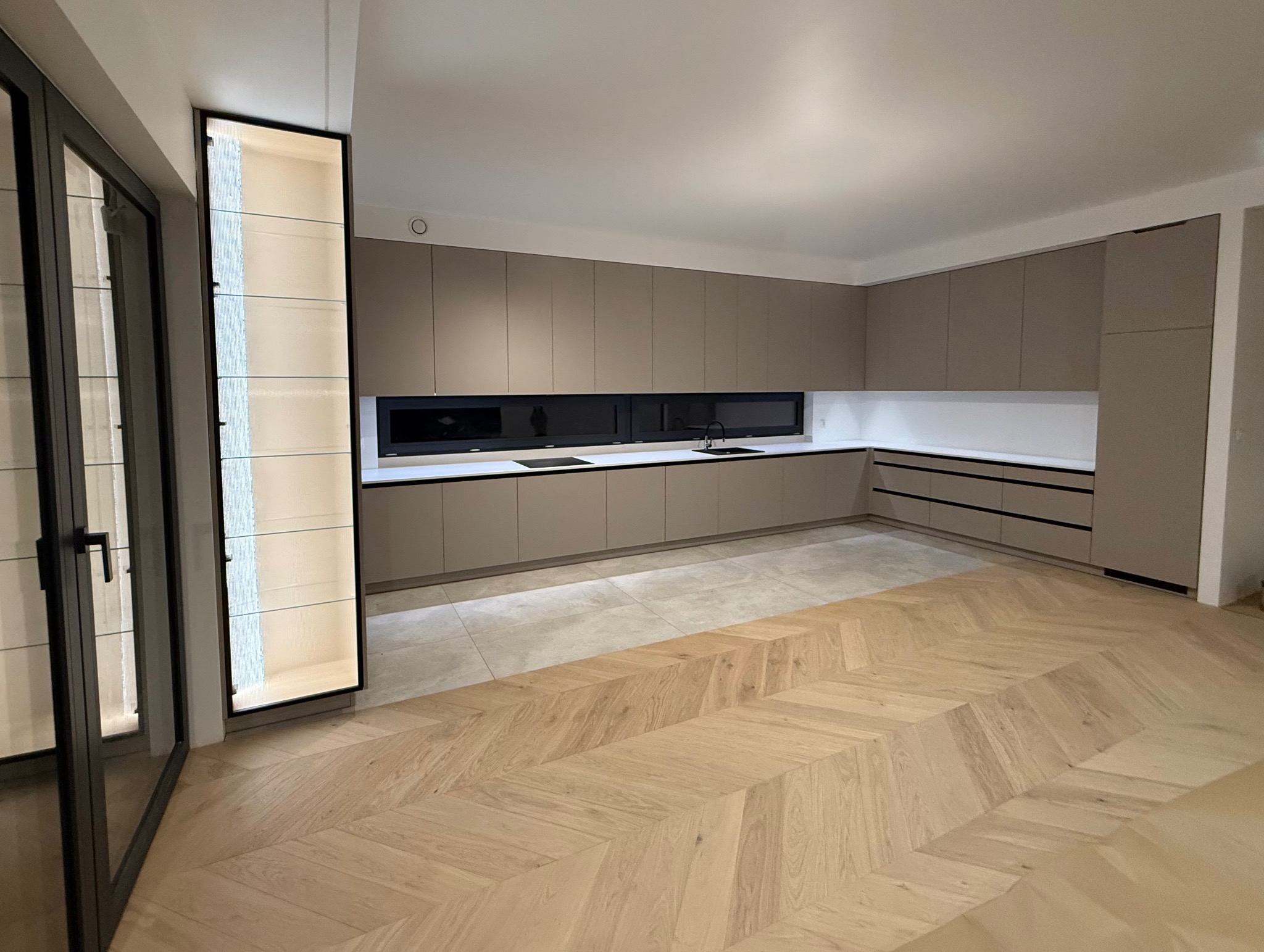 Nowoczesna kuchnia z beżowymi szafkami, czarnymi akcentami i oświetloną witryną. Parkiet w jodełkę dodaje elegancji. Minimalistyczny design z długim oknem nad blatem.