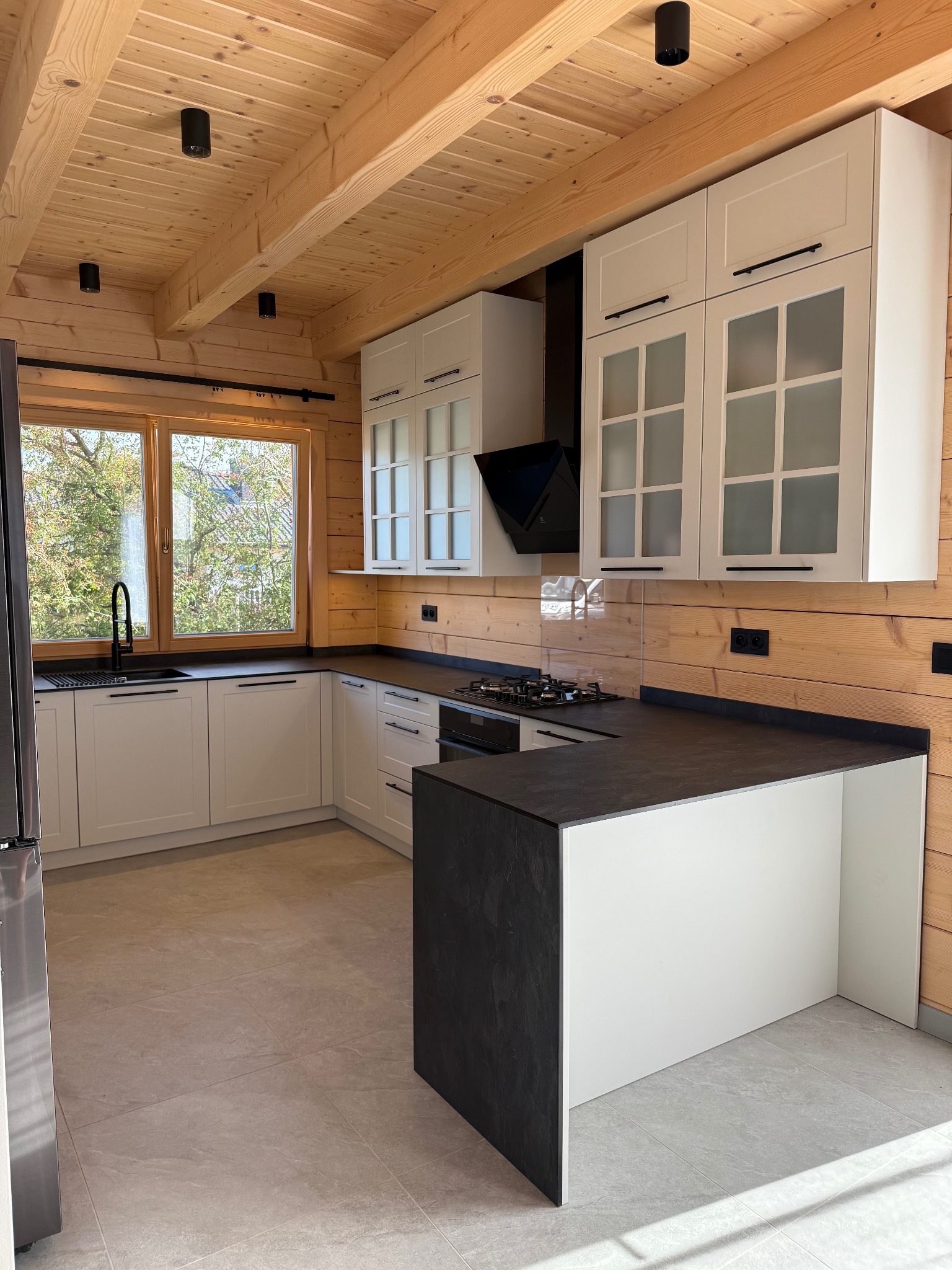 Jasna kuchnia w drewnianym domu: białe szafki z czarnymi uchwytami, ciemny blat, okno z widokiem na zieleń. Nowoczesny design w rustykalnym wnętrzu.