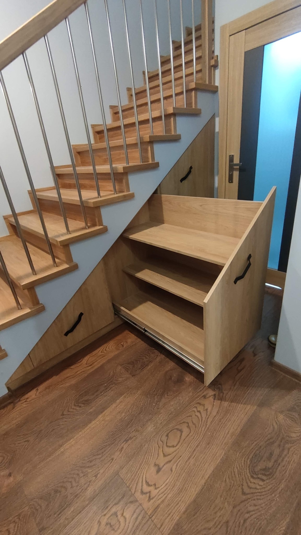 Drewniane schody z zabudową meblową pod skosami, szuflady z półkami na metalowych prowadnicach. Widoczna drewniana podłoga i fragment drzwi.