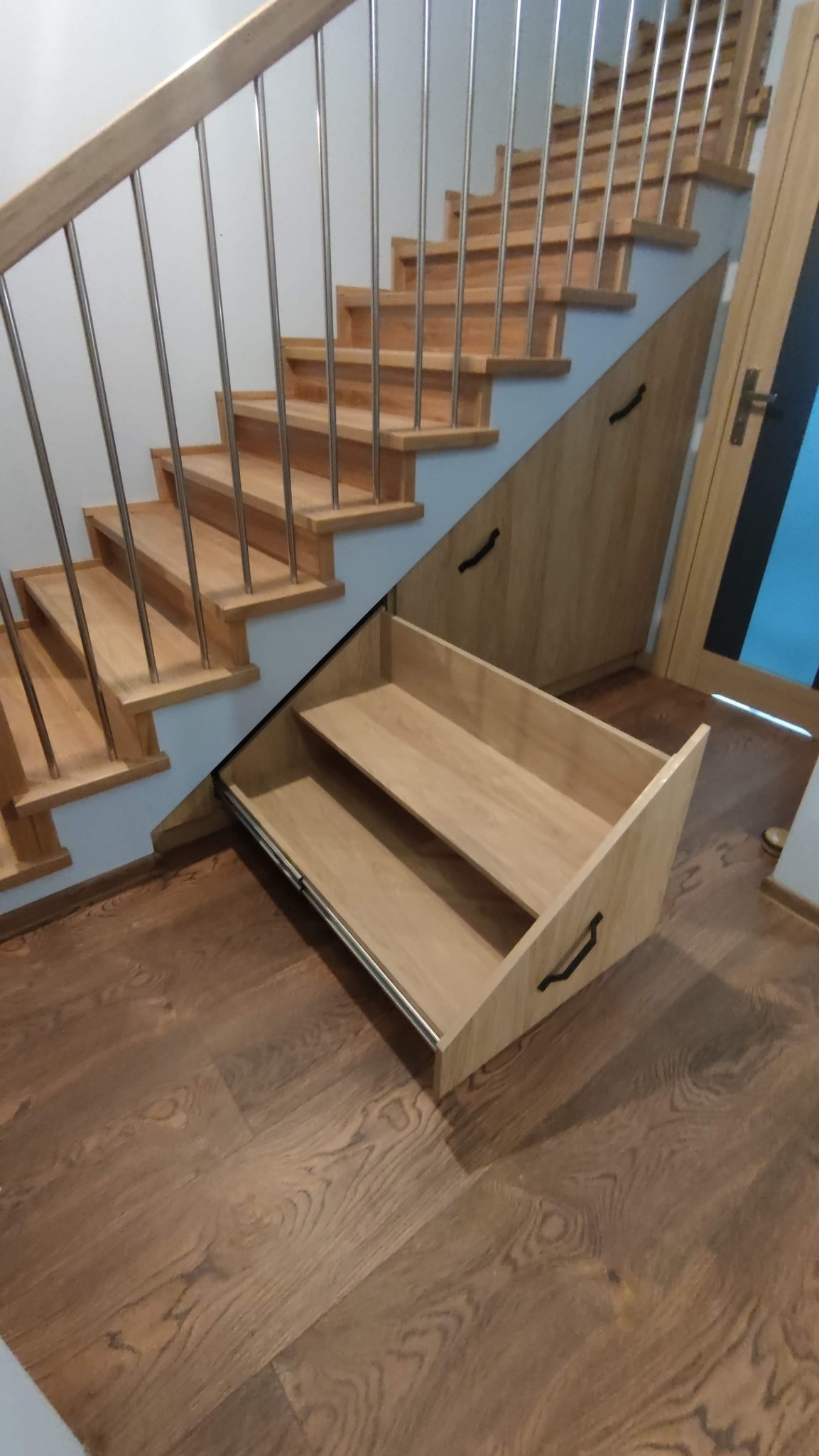 Drewniane schody z zabudową meblową pod skosami, wysuwana szuflada z dwoma półkami. Metalowe barierki, drewniana podłoga. Funkcjonalne wykorzystanie przestrzeni.