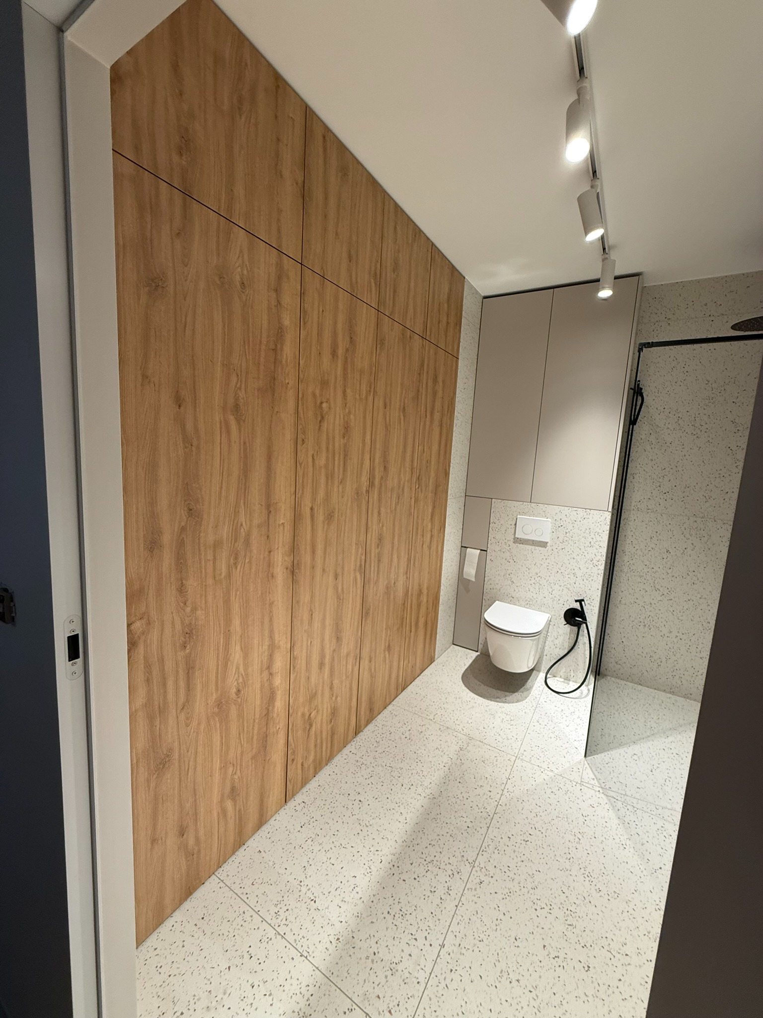 Nowoczesna łazienka z drewnianą szafą na wymiar, białą toaletą i szarą kabiną prysznicową. Minimalistyczny design, jasne oświetlenie punktowe.