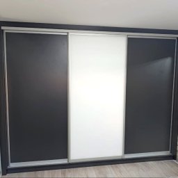 VitiukMeble - Trzydrzwiowa szafa wnękowa z przesuwnymi drzwiami: dwie czarne, jedna biała. Minimalistyczny design, srebrne obramowanie drzwi, widoczna faktura ściany w tle.