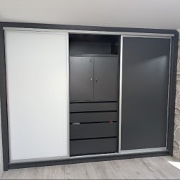 VitiukMeble - Nowoczesna szafa wnękowa z przesuwnymi drzwiami: białe skrzydło po lewej, grafitowe po prawej, centralna część z szafką i szufladami w kolorze grafitowym.