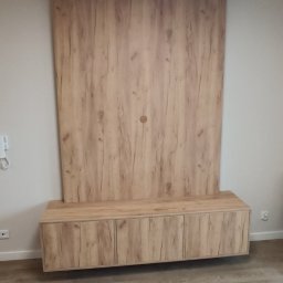 VitiukMeble - Drewniana zabudowa ściany z szafkami RTV w salonie. Fronty szafek z wyraźnym rysunkiem słojów. Minimalistyczny design, ciepłe oświetlenie sufitowe. Nowoczesne wykończenie wnętrza.