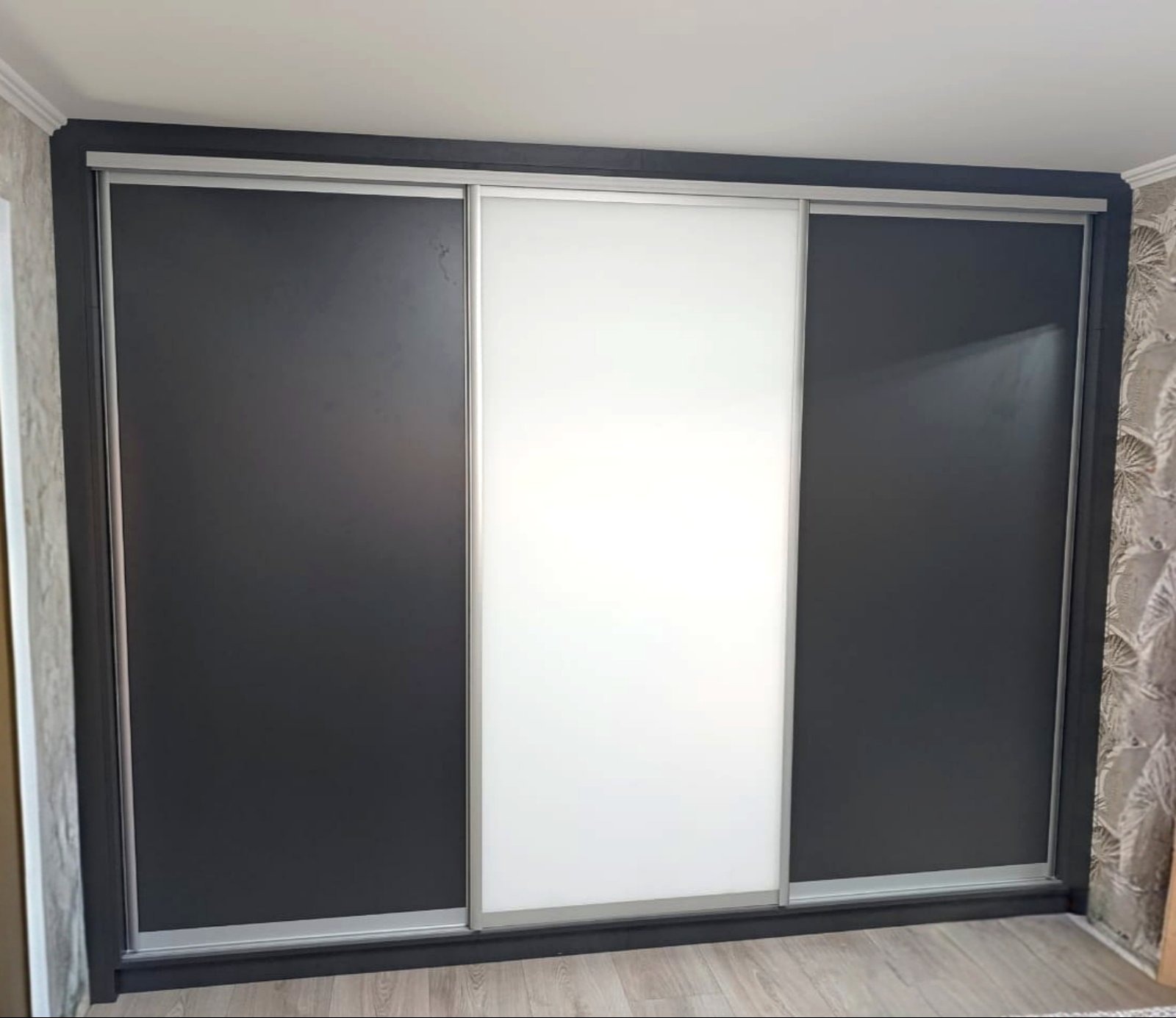 Trzydrzwiowa szafa wnękowa z przesuwnymi drzwiami: dwie czarne, jedna biała. Minimalistyczny design, srebrne obramowanie drzwi, widoczna faktura ściany w tle.