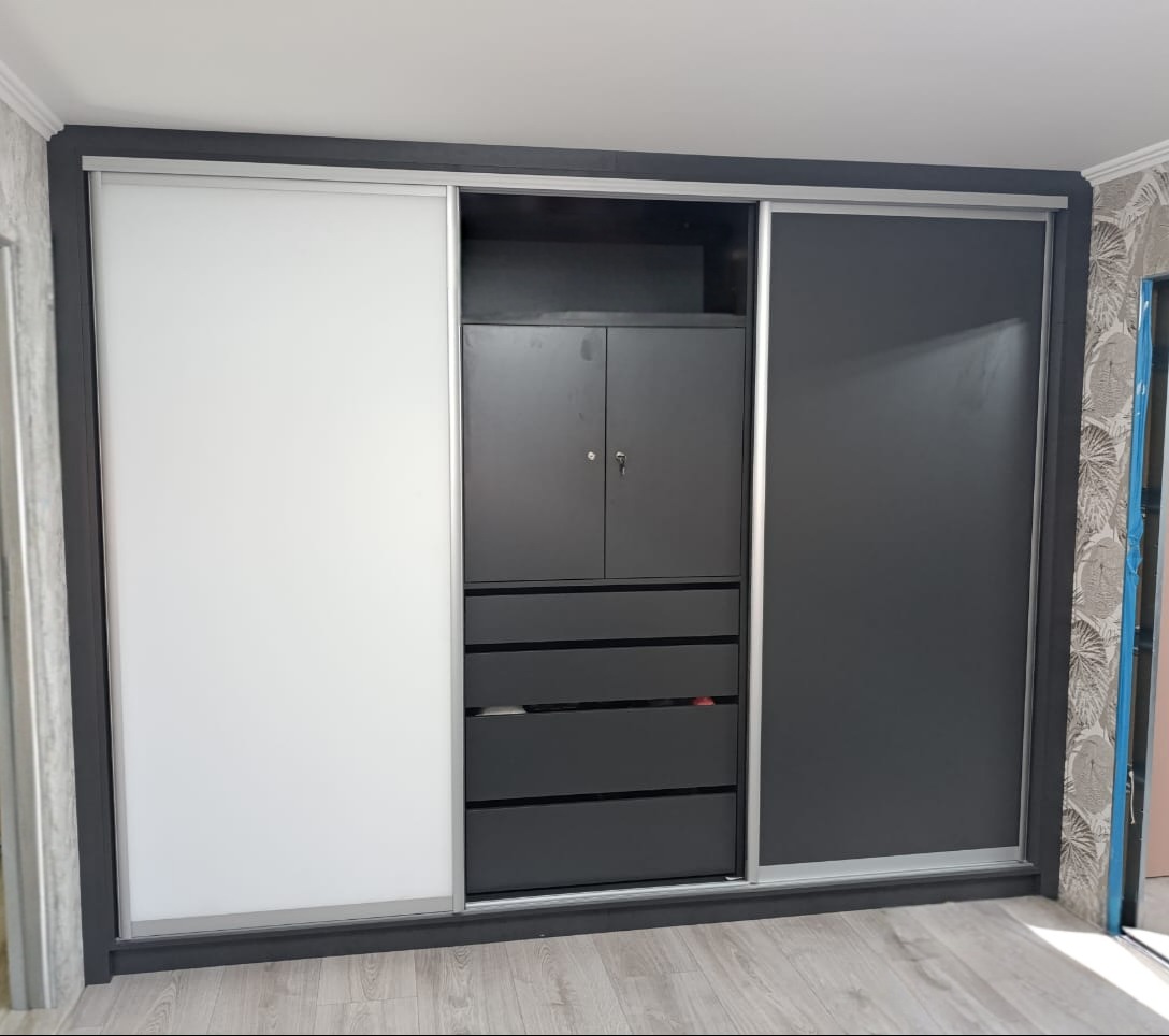 Nowoczesna szafa wnękowa z przesuwnymi drzwiami: białe skrzydło po lewej, grafitowe po prawej, centralna część z szafką i szufladami w kolorze grafitowym.