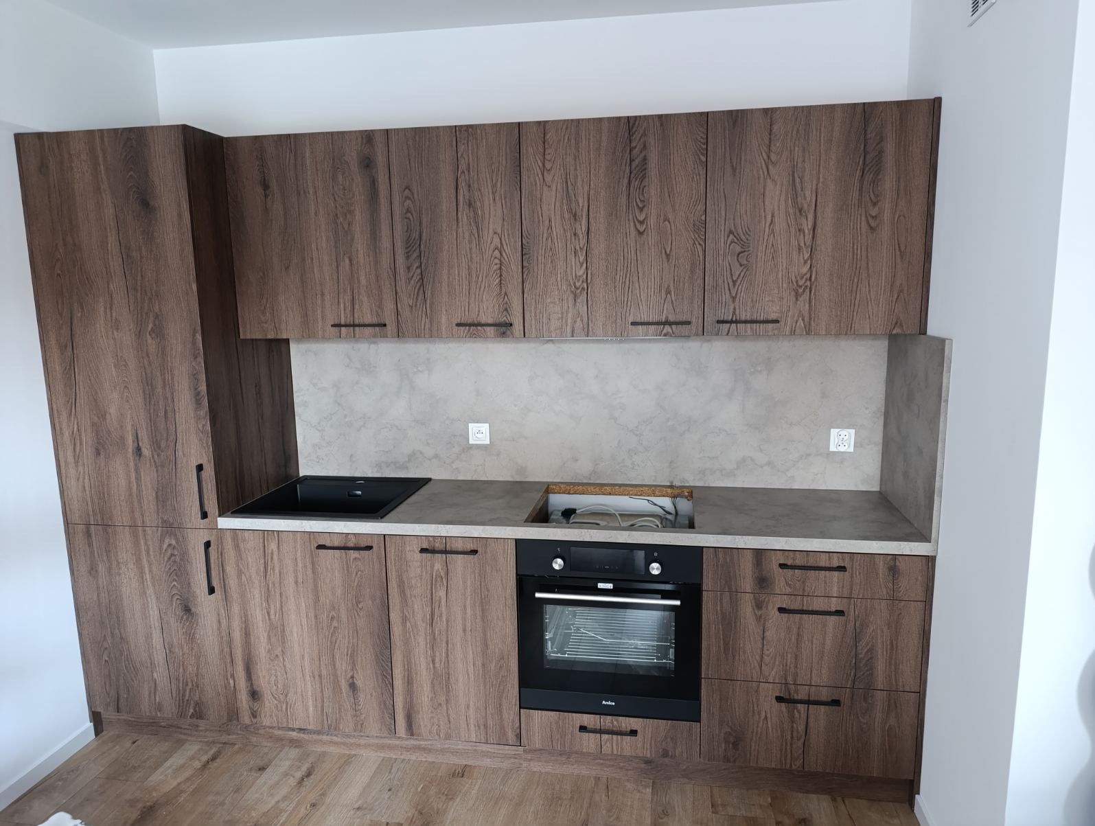 Nowoczesna kuchnia z drewnianymi frontami i czarnym zlewem, oświetlona naturalnym światłem. Widoczne gniazdka elektryczne na ścianie nad blatem.