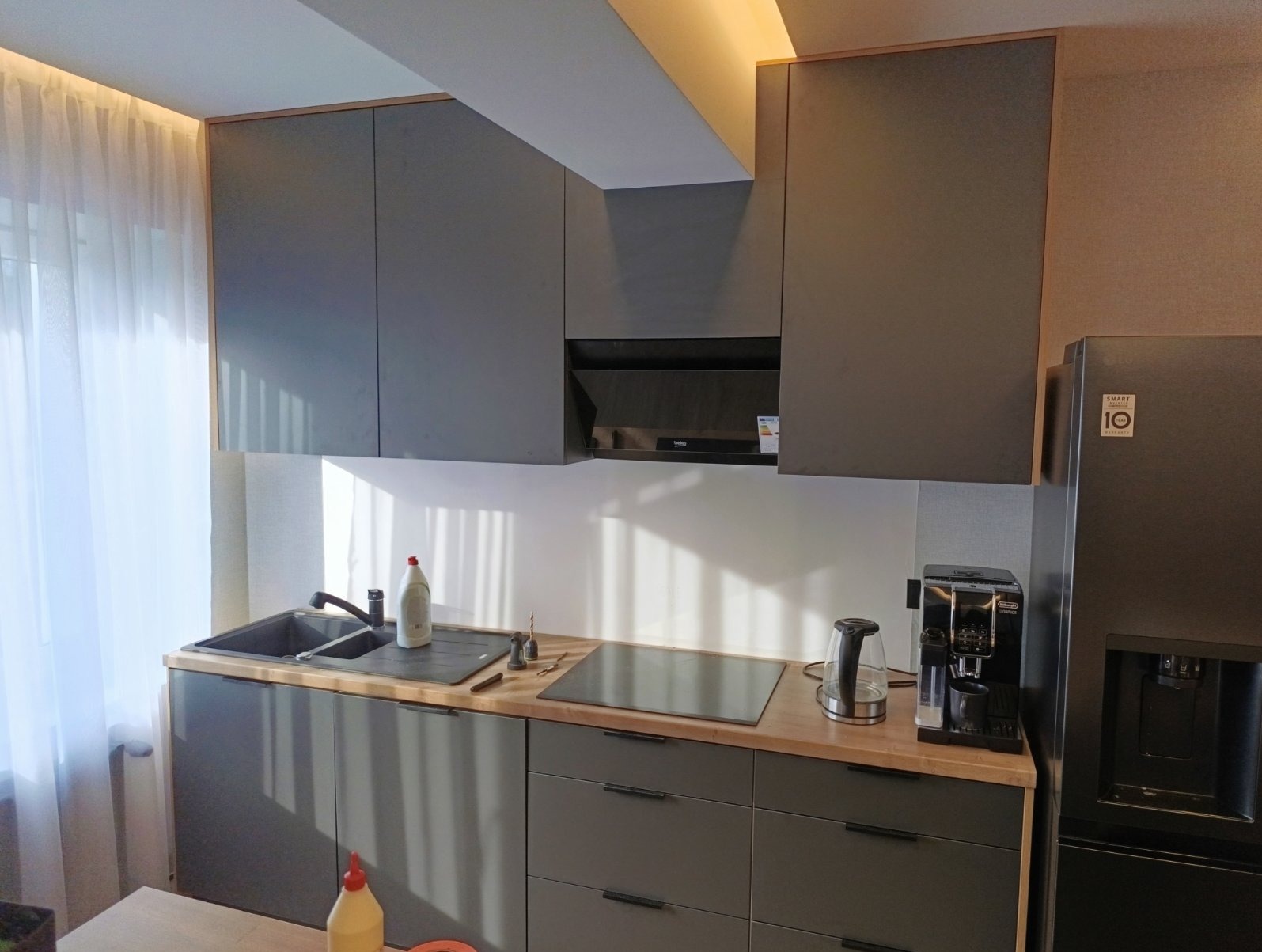 Nowoczesna kuchnia z szarymi szafkami, drewnianym blatem i sprzętem AGD. Widoczne okno z firanką i promienie słoneczne. Minimalistyczny design z dbałością o detale.