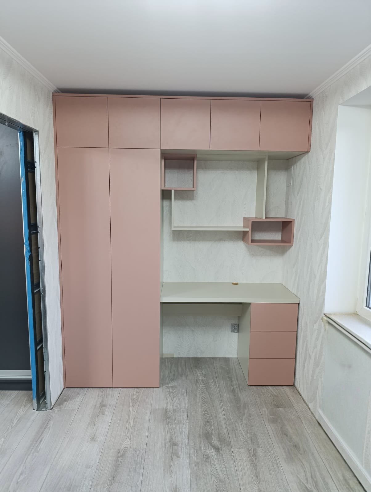 Różowa zabudowa meblowa z szafą i biurkiem w pokoju. Szafki wiszące nad blatem biurka z półkami. Nowoczesny design, jasne wnętrze, panele podłogowe.