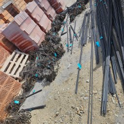 ETER STAL Adrian Pasiniewicz - Cegły Termoton na palecie i wiązki prętów zbrojeniowych z niebieskimi etykietami, ułożone na ziemi. Widok z góry, ukośny kadr.