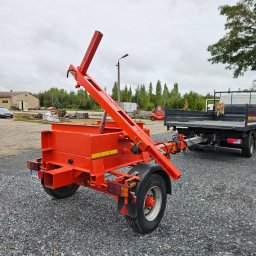 ETER STAL Adrian Pasiniewicz - Czerwona przyczepa z podnośnikiem hydraulicznym, zaparkowana na żwirowym placu, podpięta do ciężarówki. W tle budynki magazynowe i drzewa.