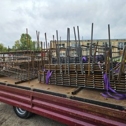 ETER STAL Adrian Pasiniewicz - Transport zbrojenia na pace ciężarówki, zabezpieczone fioletowymi pasami. Widoczne elementy konstrukcyjne i tło z budynkiem. Ujęcie z boku, pochmurne niebo.