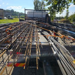 ETER STAL Adrian Pasiniewicz - Transport stali zbrojeniowej na budowę. Widok z góry na ciężarówkę wypełnioną elementami zbrojeniowymi, zabezpieczonymi pomarańczowymi pasami. Słoneczny dzień.