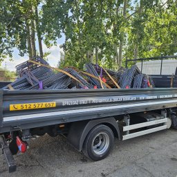 ETER STAL Adrian Pasiniewicz - Ciężarówka z platformą załadowaną stalą zbrojeniową, zabezpieczoną taśmami. Widoczne drzewa w tle i numer telefonu na burcie pojazdu.