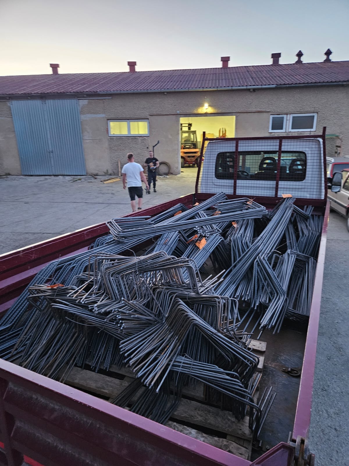 Stalowe pręty zbrojeniowe w kształcie litery U, ułożone w skrzyni samochodu dostawczego na tle budynku magazynowego z otwartą bramą i wózkiem widłowym w środku.