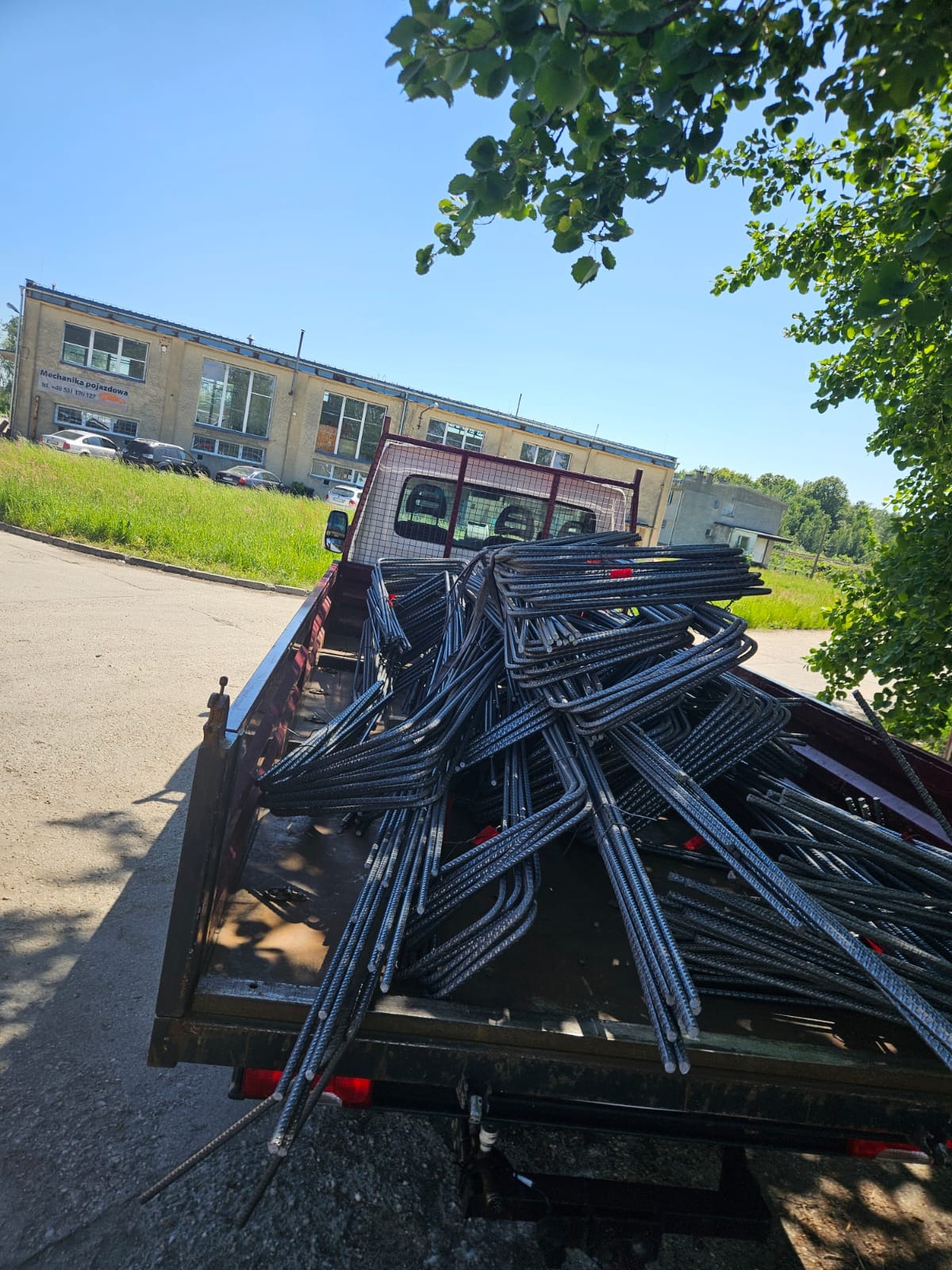 Transport zbrojenia w kształcie litery U na pace samochodu dostawczego. W tle budynek z napisem 'Mechanika pojazdowa' i zieleń.