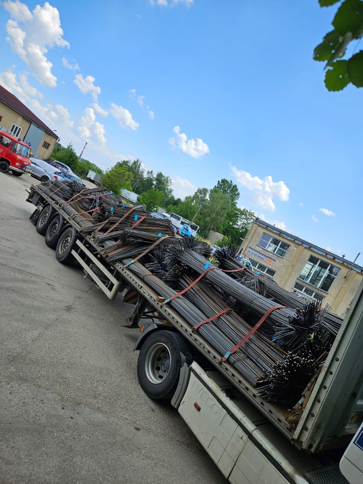 Ciężarówka z ładunkiem prętów zbrojeniowych, zabezpieczonych czerwonymi pasami, na tle błękitnego nieba z chmurami i budynków. Widok z boku, lekki kąt.