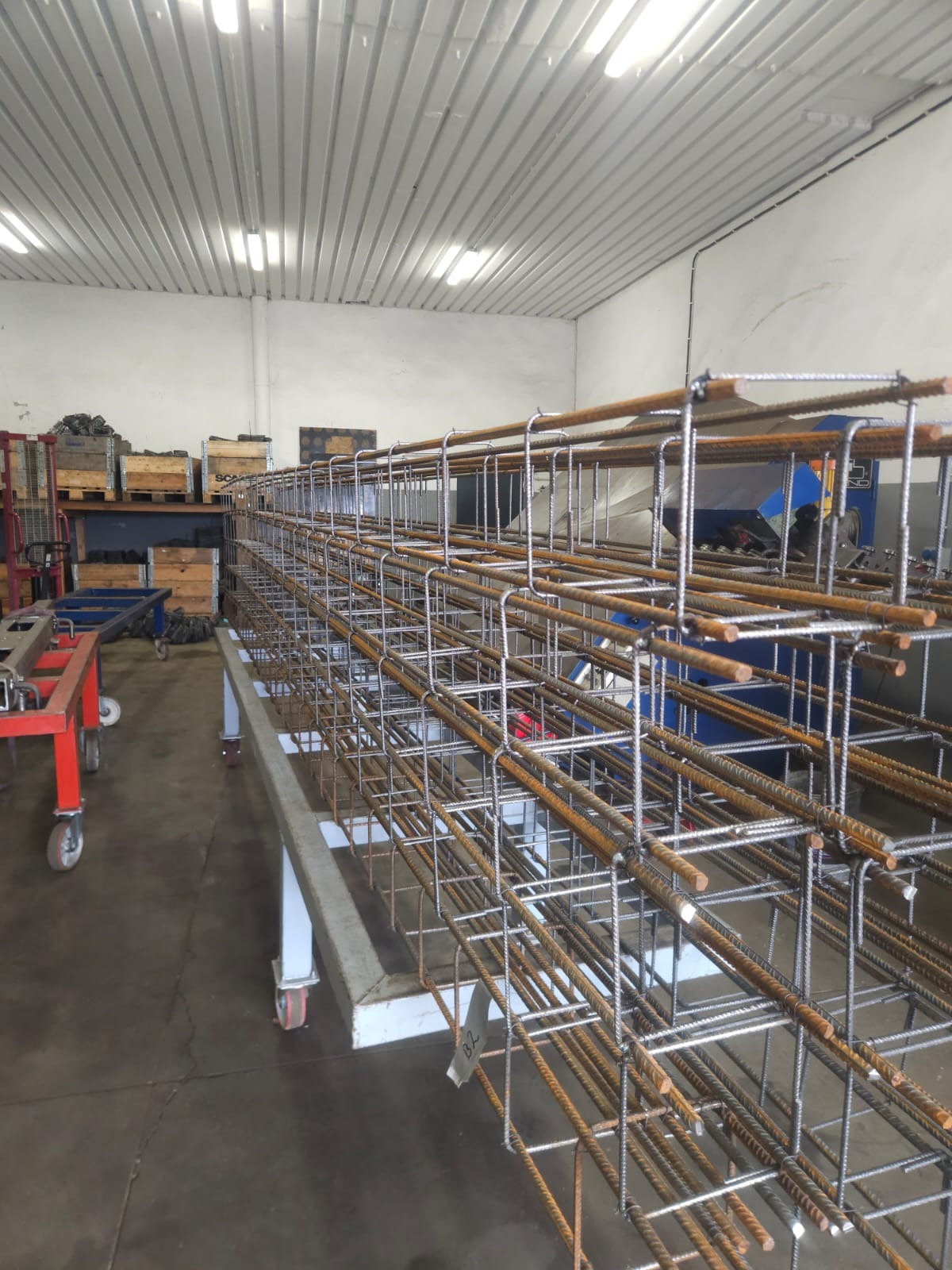 Gotowa klatka zbrojeniowa na metalowym wózku w hali produkcyjnej. Widoczne elementy konstrukcyjne i spawane łączenia. W tle maszyny i materiały budowlane.
