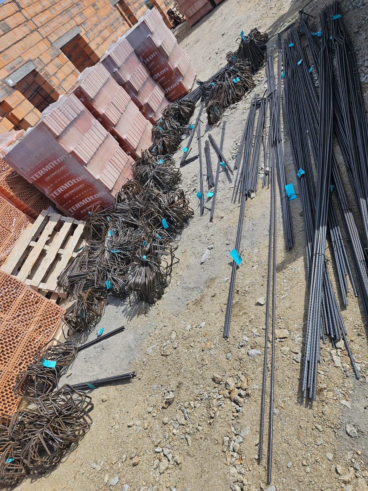 Cegły Termoton na palecie i wiązki prętów zbrojeniowych z niebieskimi etykietami, ułożone na ziemi. Widok z góry, ukośny kadr.