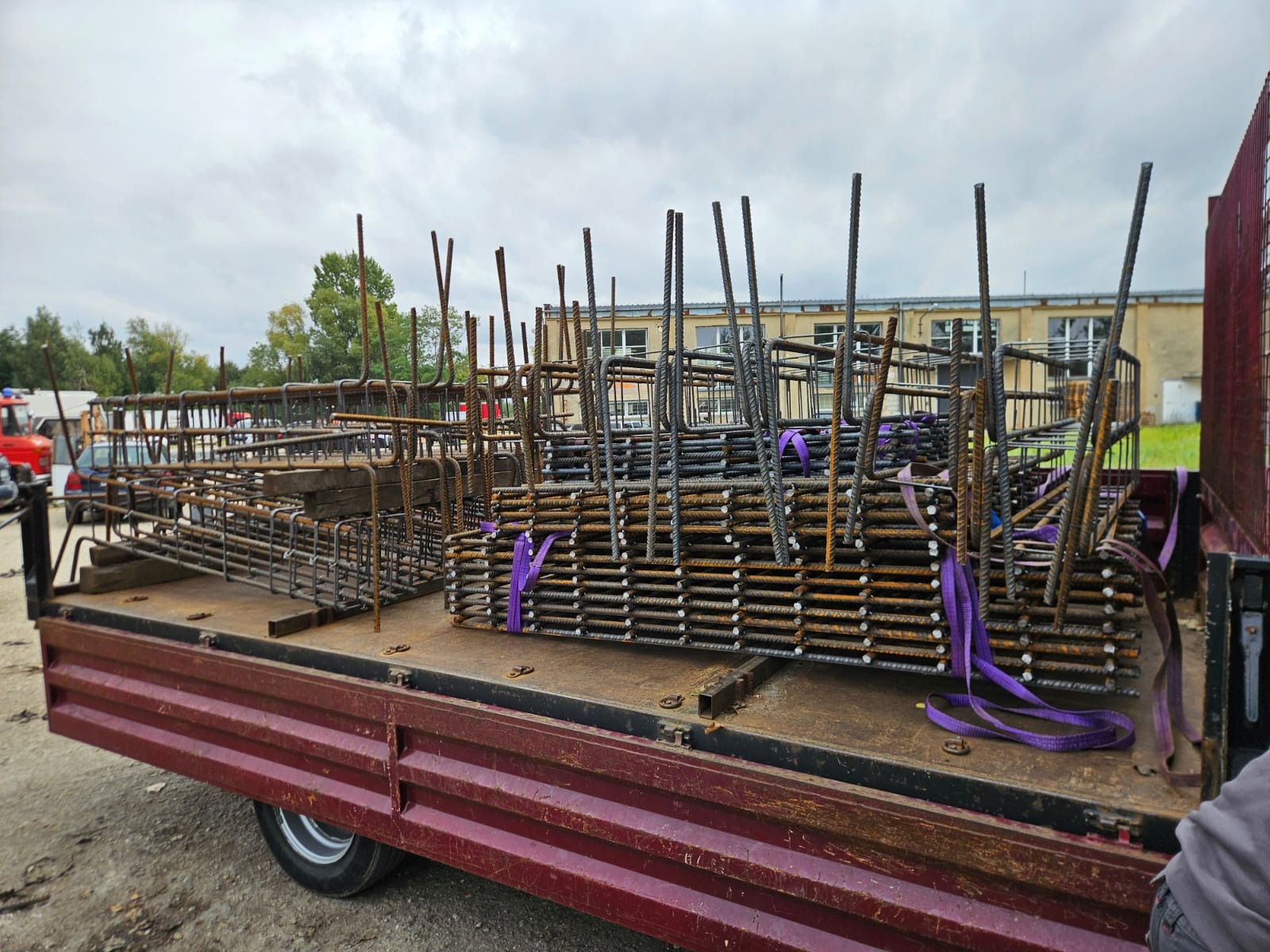 Transport zbrojenia na pace ciężarówki, zabezpieczone fioletowymi pasami. Widoczne elementy konstrukcyjne i tło z budynkiem. Ujęcie z boku, pochmurne niebo.