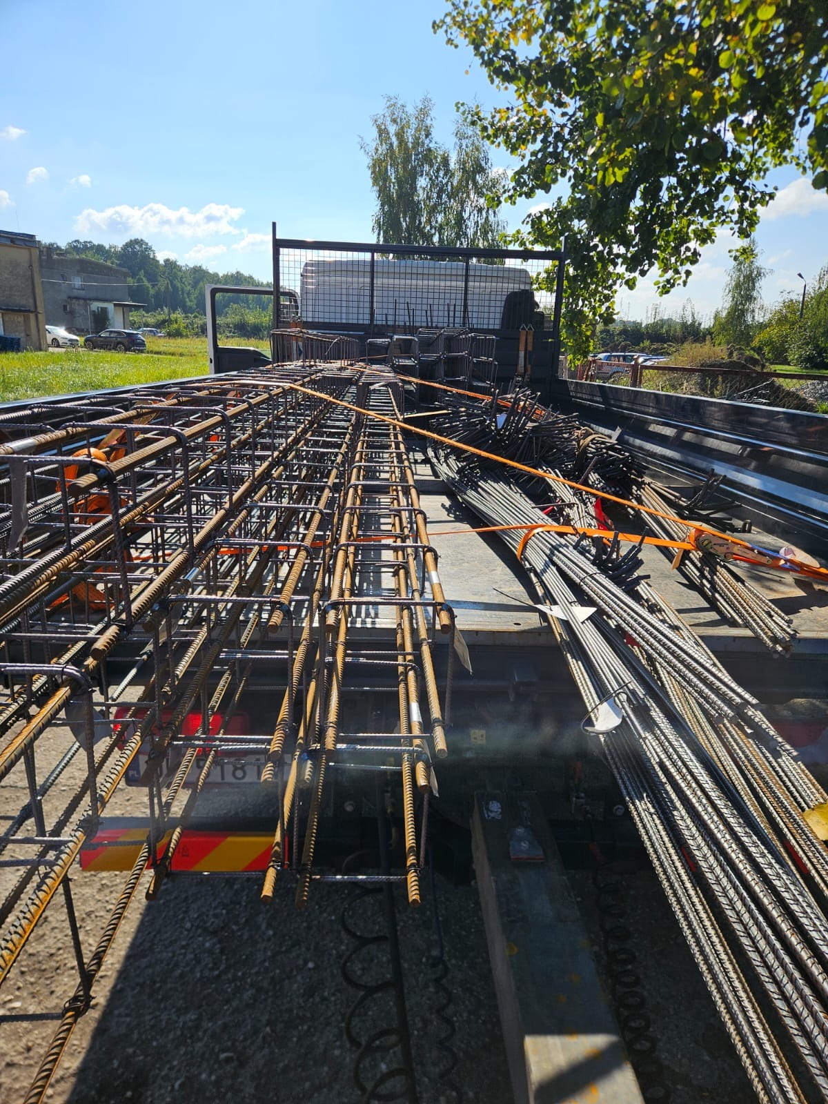 Transport stali zbrojeniowej na budowę. Widok z góry na ciężarówkę wypełnioną elementami zbrojeniowymi, zabezpieczonymi pomarańczowymi pasami. Słoneczny dzień.