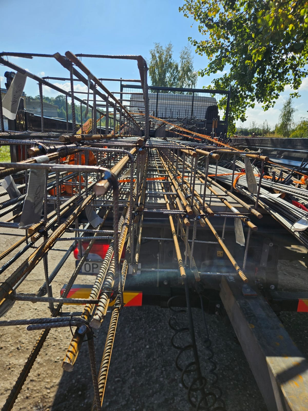 Transport zbrojenia na przyczepie w słoneczny dzień. Widoczne elementy konstrukcji i tablica rejestracyjna pojazdu. Zastosowanie stali żebrowanej.
