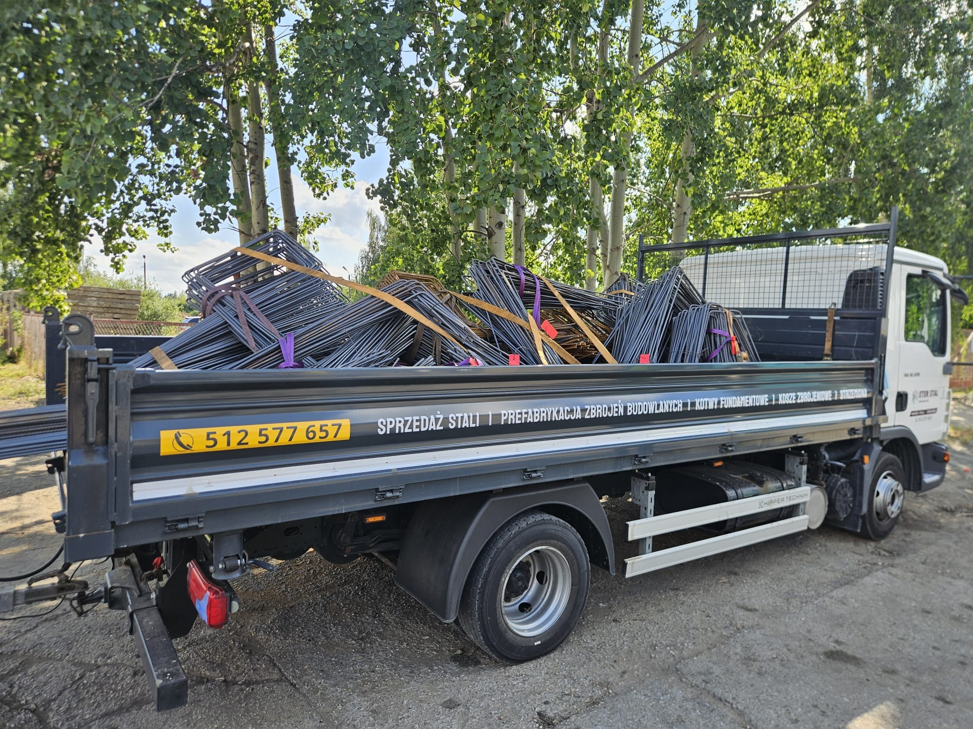 Ciężarówka z platformą załadowaną stalą zbrojeniową, zabezpieczoną taśmami. Widoczne drzewa w tle i numer telefonu na burcie pojazdu.