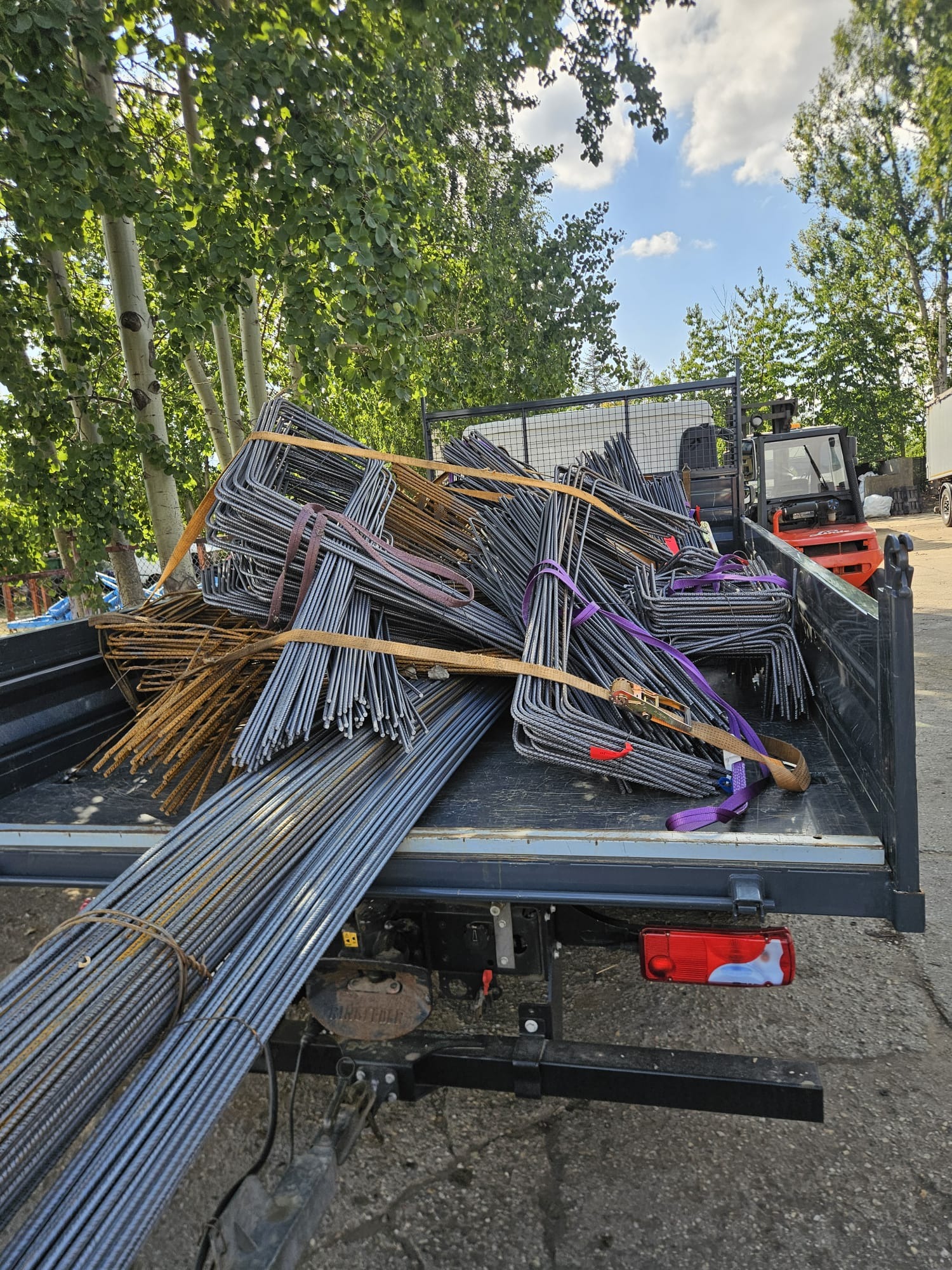 Stal zbrojeniowa w transporcie: pręty i strzemiona zabezpieczone pasami w skrzyni ładunkowej samochodu dostawczego, w tle wózek widłowy i drzewa.