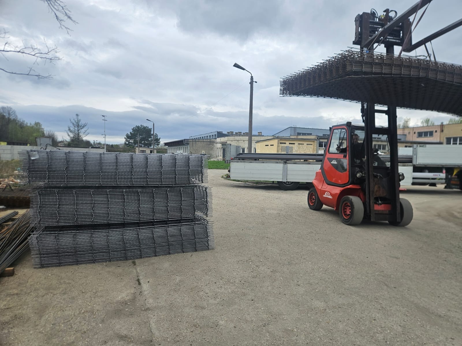 Czerwony wózek widłowy Linde transportuje siatkę zbrojeniową na placu budowy. W tle budynki i zaparkowane przyczepy. Pochmurne niebo.