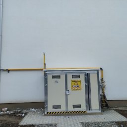 SPAWMAKS Andrii Maksimchuk - Zewnętrzna szafka gazowa z żółtą instalacją przy budynku. Widoczna drabina oparta o szafkę i fragment chodnika z kostki brukowej. Osoba w tle.