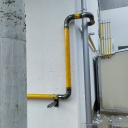 SPAWMAKS Andrii Maksimchuk - Żółta instalacja gazowa na białej ścianie budynku, widoczne spawane łączenia rur i elementy mocujące. Rura spustowa obok. Fragment chodnika z kostki brukowej.
