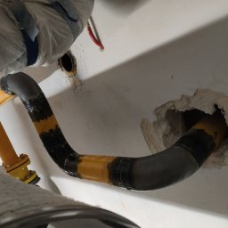 SPAWMAKS Andrii Maksimchuk - Żółto-czarna rura gazowa, częściowo zaizolowana, przechodzi przez otwór w ścianie. Widoczne łączenia i elementy instalacji. Ujęcie z bliska, z dołu.