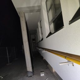 SPAWMAKS Andrii Maksimchuk - Zewnętrzny korytarz budynku w nocnej scenerii. Widoczny betonowy filar, żółta poręcz i okna. W tle siatka ogrodzeniowa i drabina.