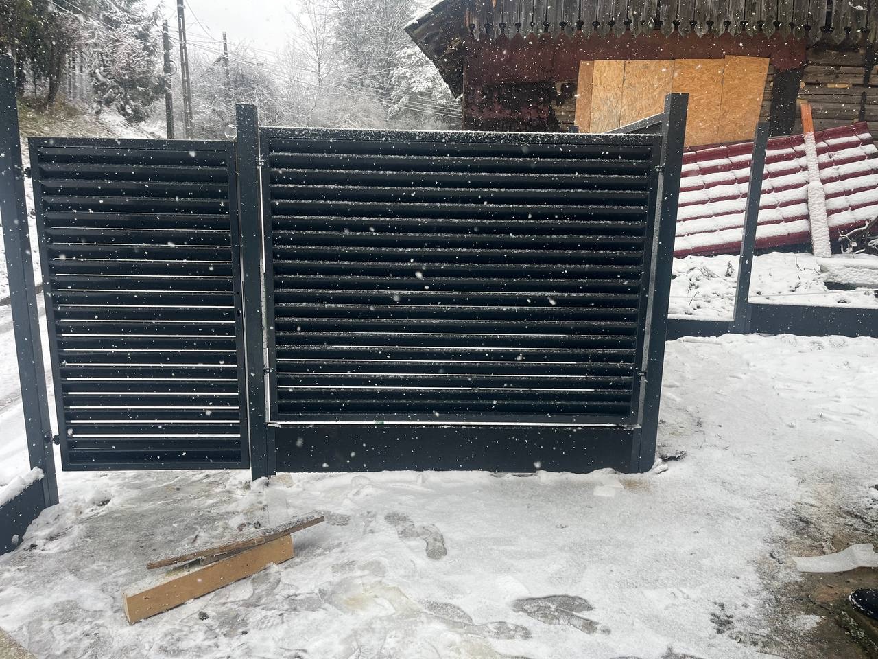 Nowoczesna, czarna brama lamelowa z metalu, zamontowana w zimowej scenerii. Widoczne opady śniegu i fragmenty drewnianego budynku w tle. Minimalistyczny design.