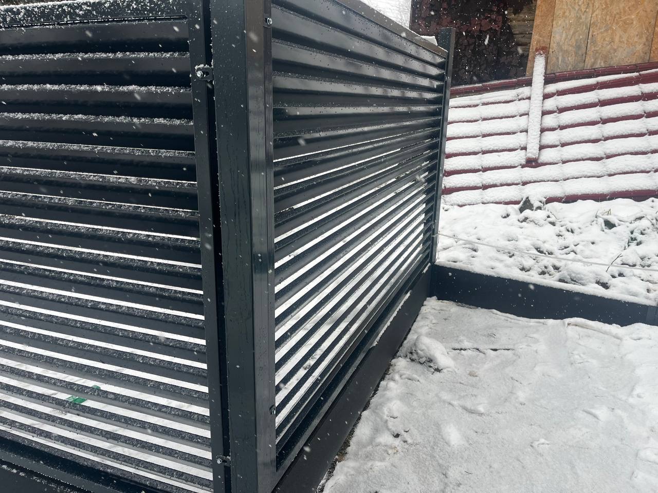 Nowoczesne, poziome ogrodzenie panelowe w kolorze antracytowym, pokryte śniegiem. Widoczne detale konstrukcyjne i fragment otoczenia w zimowej scenerii.