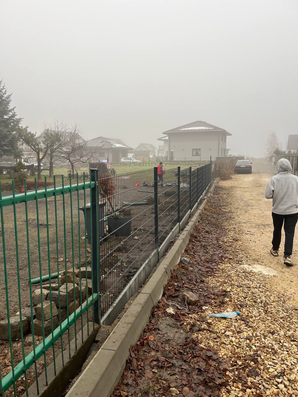 Montaż ogrodzenia panelowego na posesji w mglisty dzień. Widoczny fragment starego ogrodzenia i osoba w oddali przy nowym ogrodzeniu.