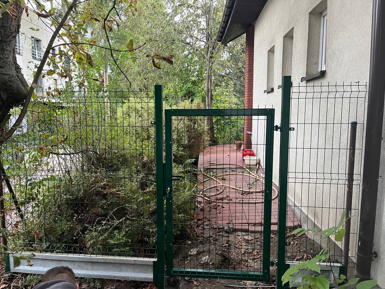 Zielone ogrodzenie panelowe z furtką, zamontowane przy budynku z ceglanym kominem. Widoczne krzewy i drzewa w tle. Częściowo widoczna osoba w dolnym lewym rogu.