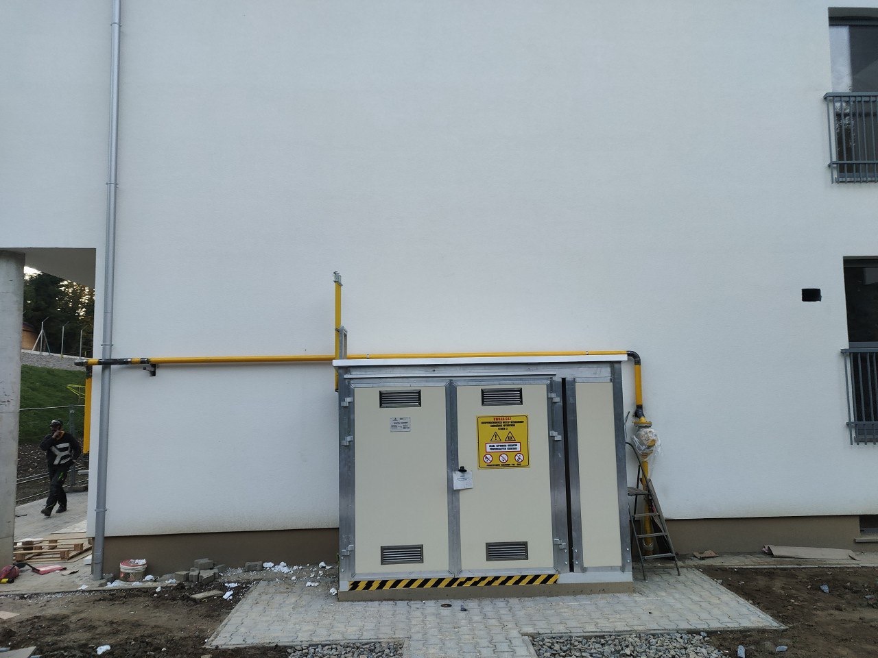 Zewnętrzna szafka gazowa z żółtą instalacją przy budynku. Widoczna drabina oparta o szafkę i fragment chodnika z kostki brukowej. Osoba w tle.