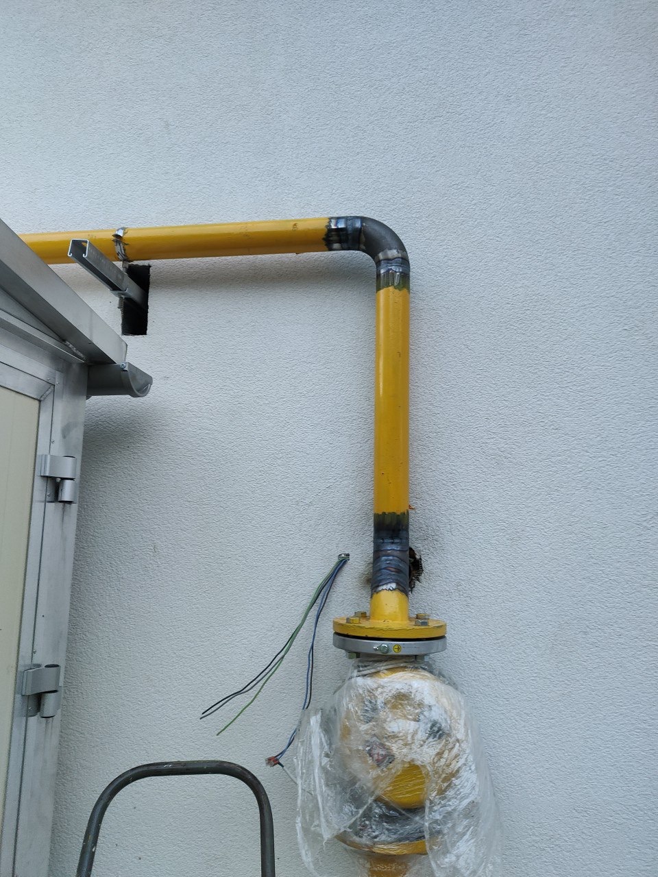 Żółta instalacja gazowa na zewnątrz budynku, z widocznymi spawami i elementami mocującymi. Część instalacji owinięta folią ochronną, z przewodami obok.
