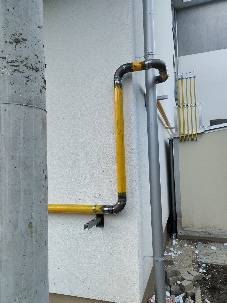 Żółta instalacja gazowa na białej ścianie budynku, widoczne spawane łączenia rur i elementy mocujące. Rura spustowa obok. Fragment chodnika z kostki brukowej.